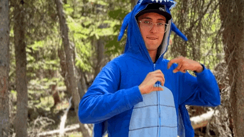 a man wearing a blue hoodie with a shark hood and ears is standing in the woods .