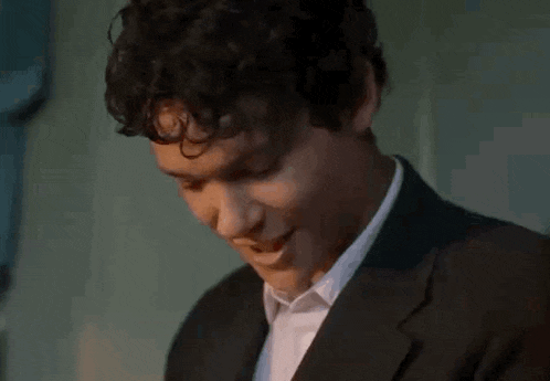 a young man with curly hair is wearing a suit and white shirt and looking down .
