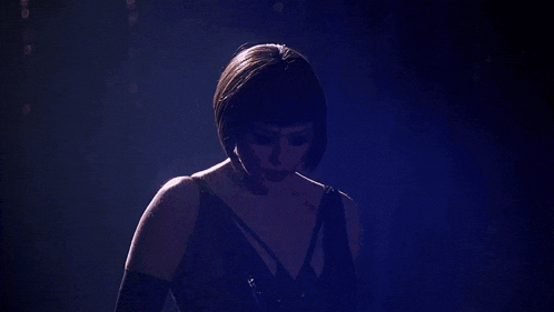 a woman in a black dress is standing in a dark room looking at the camera