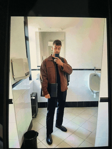 a man taking a selfie in a bathroom with a urinal in the background