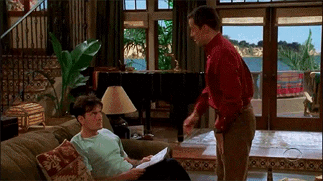 two men are sitting on a couch in a living room talking to each other