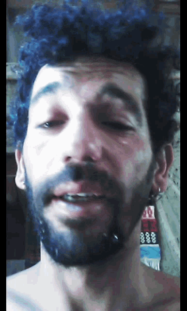a man with blue hair and a beard looks at the camera with his eyes closed