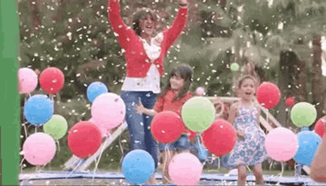a woman and two girls are standing on a trampoline with balloons falling from the sky .