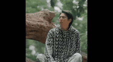 a man is sitting on a tree branch in the woods wearing a sweater .