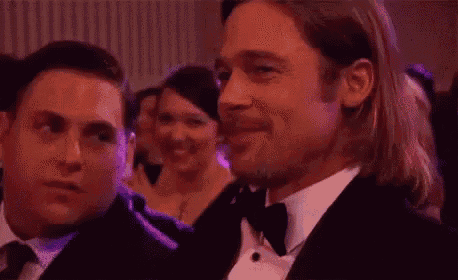 a man in a tuxedo and tie is smiling while sitting next to another man in a crowd .