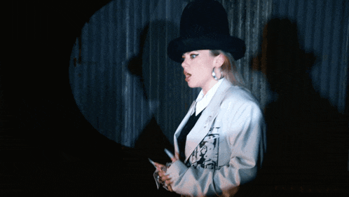 a woman wearing a black top hat and a white jacket stands in front of a metal wall