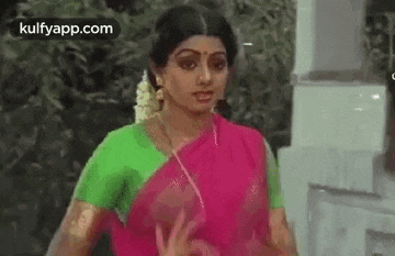a woman in a pink saree and green blouse is standing in front of a white building .