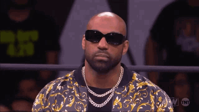 a bald man wearing sunglasses and a gold chain looks at the camera