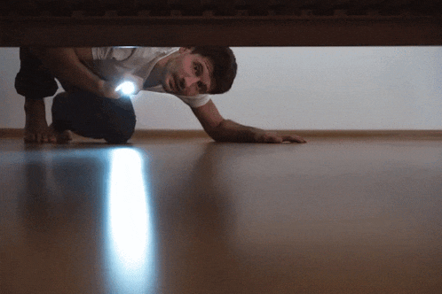 a man crawls under a bed holding a flashlight