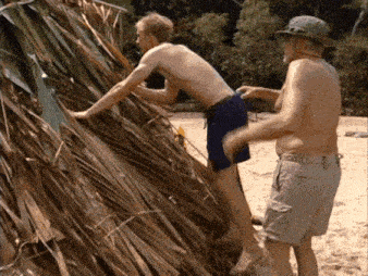 a shirtless man climbs a pile of branches while another man looks on