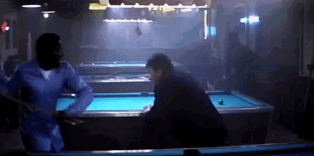 two men are playing pool in a dark room and one of them is wearing a blue shirt