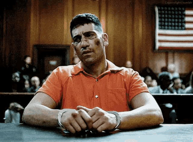 a man in an orange shirt is sitting at a table with his hands in handcuffs in front of an american flag .