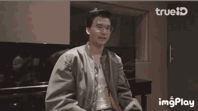 a man wearing glasses and a bomber jacket is clapping his hands in front of a piano in a room with a trueid logo