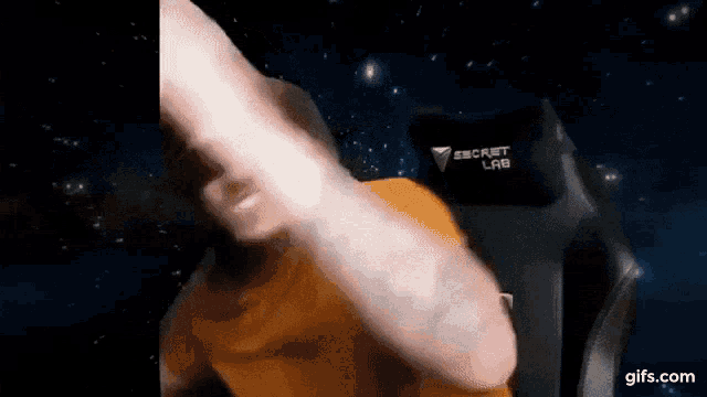 a man in an orange shirt is giving a thumbs up in front of a secret lab chair