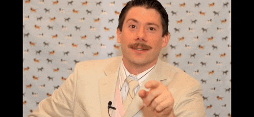 a man with a mustache wearing a suit and tie points at the camera