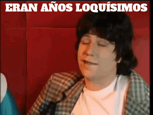 a man in a plaid jacket is sitting in front of a red wall with the words " eran anos loquisimos " above him