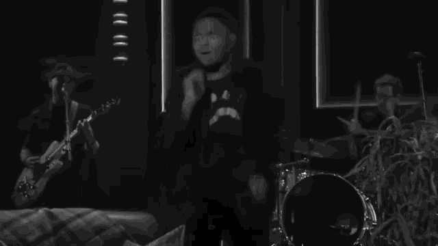 a black and white photo of a man singing and dancing in front of a drum set .