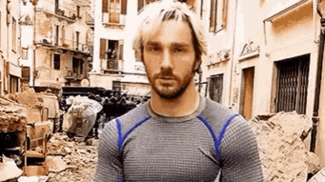 a man with blonde hair and a beard is standing in a destroyed city .