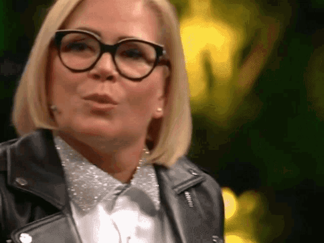 a woman wearing glasses and a leather jacket looks at the camera