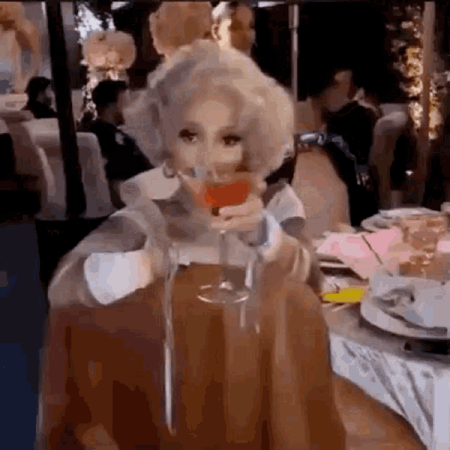 a woman in a wig is sitting at a table drinking a glass of wine .