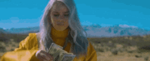 billie eilish is wearing a yellow jacket and holding a bunch of money in a desert .