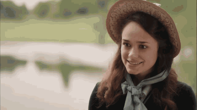 a woman wearing a hat and a scarf smiles in front of a body of water