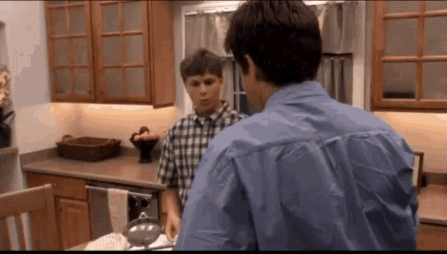 a man in a blue shirt is talking to another man in a plaid shirt in a kitchen