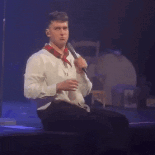 a man is sitting on a stage holding a microphone and making a funny face .