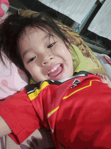 a little girl wearing a red shirt with a yellow stripe on the sleeves is smiling