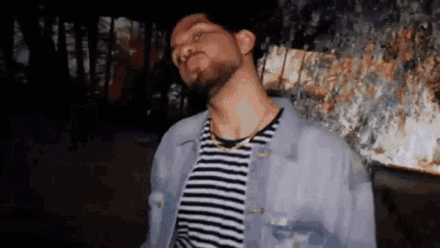 a man with a beard wearing a striped shirt and a denim jacket is standing in front of a fire .
