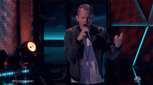 a man singing into a microphone on a stage with a nbc logo behind him