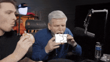 two men are eating donuts in front of a microphone and one of them is holding a piece of paper with the letter s on it