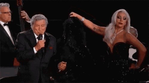 a woman in a black dress is standing next to a man in a tuxedo while singing into a microphone .
