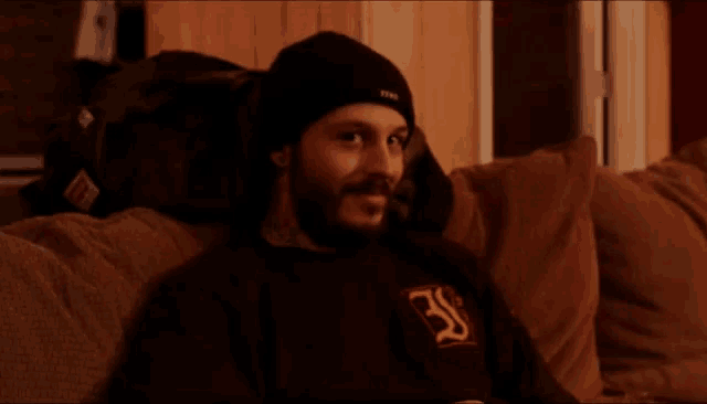 a man is sitting on a couch wearing a black shirt with the letter s on it