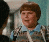 a young boy wearing suspenders and a blue shirt is talking to another person .