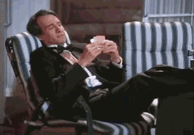 a man in a tuxedo and tie is sitting in a chair holding a pair of playing cards .