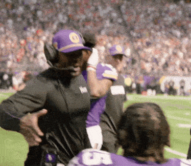 a man wearing headphones and a purple hat is standing on a football field with players .