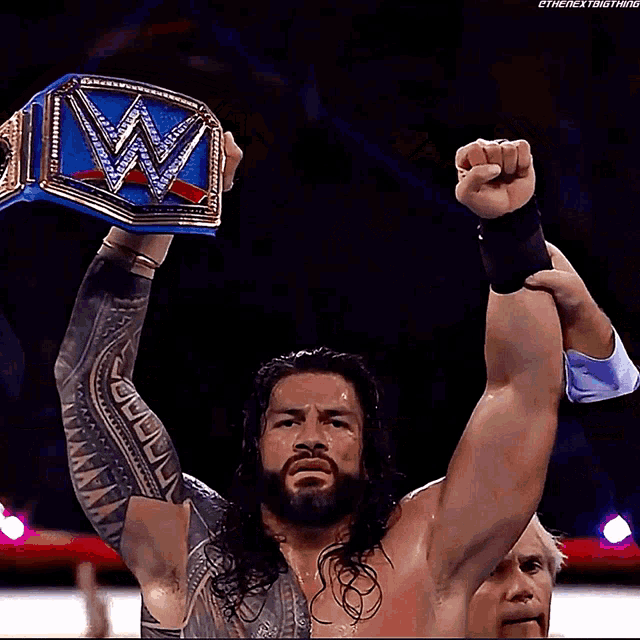 roman reigns is holding a blue wrestling championship belt in the air