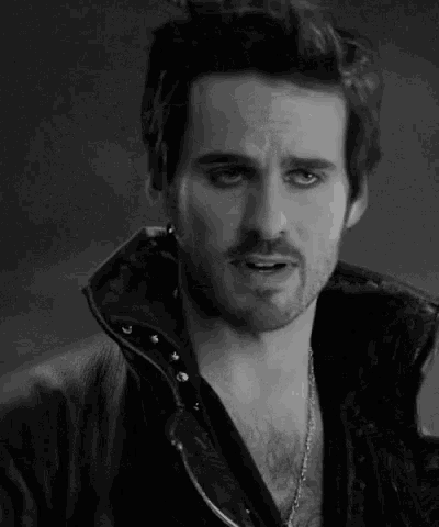 a black and white photo of a man wearing a leather jacket and necklace .