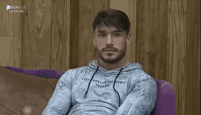 a man with a beard is sitting on a couch wearing a grey hoodie .