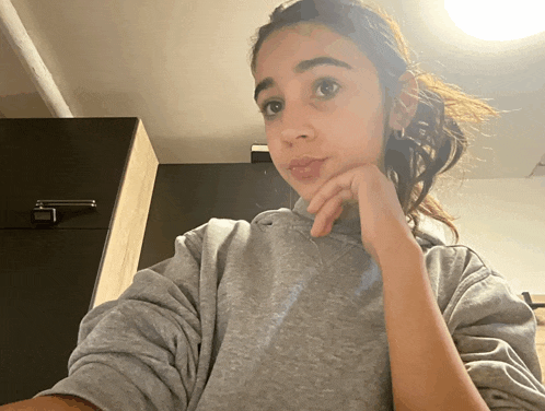 a girl wearing a grey sweatshirt looks at the camera with her hand on her chin
