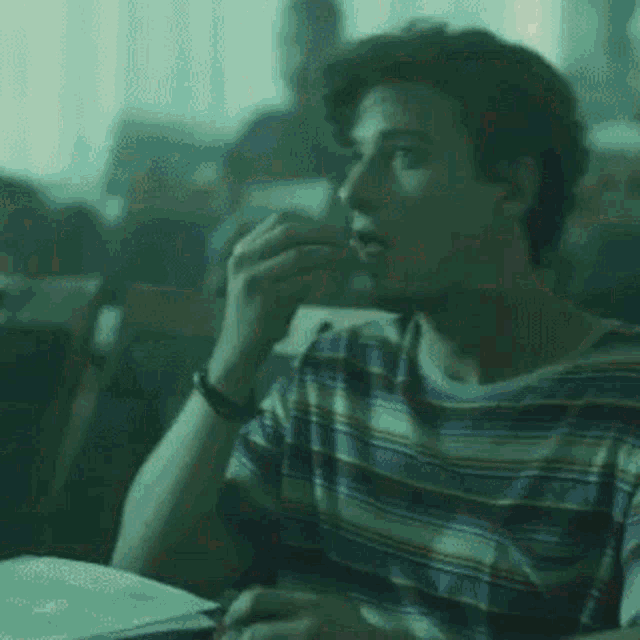 a young man in a striped shirt is sitting at a table in a classroom eating something .