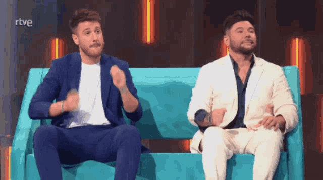 two men in suits are sitting on a blue couch with rtve written on the bottom