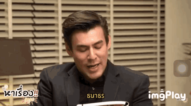 a man in a suit is sitting in front of a window with blinds and the word imgplay is on the bottom of the screen