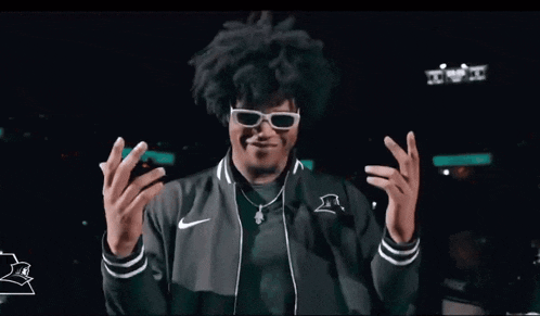 a man wearing sunglasses and a nike jacket is laughing and holding a cell phone .
