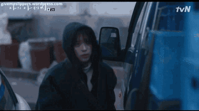 a woman in a hooded jacket is standing in front of a blue truck with tvn written on the bottom right