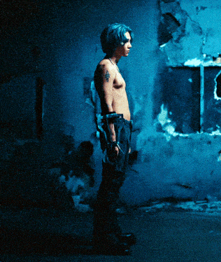 a shirtless man with blue hair and a tattoo on his arm stands in front of a blue wall