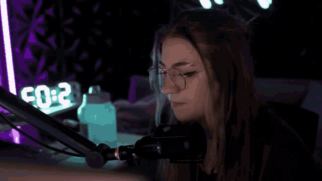 a woman wearing glasses stands in front of a microphone in front of a purple clock that says 2:02