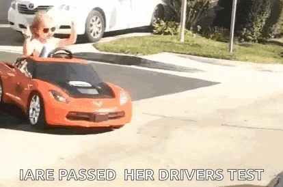 a little girl is driving a red toy car on a street .