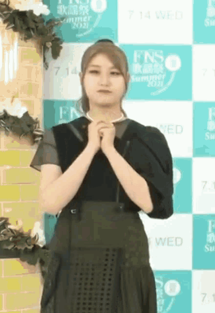 a woman in a black dress is standing in front of a wall with flowers and a sign that says fns summer 2021 .